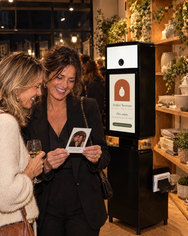 Lors de l'inauguration d'une boutique de c&eacute;ramique, deux amies rient en regardant leur tirage &agrave; fen&ecirc;tre en arche ; derri&egrave;re elles, l'&eacute;cran de la borne affiche une arche terre cuite et &laquo; Pi&egrave;ce unique &raquo;. Mise en situation g&eacute;n&eacute;r&eacute;e.