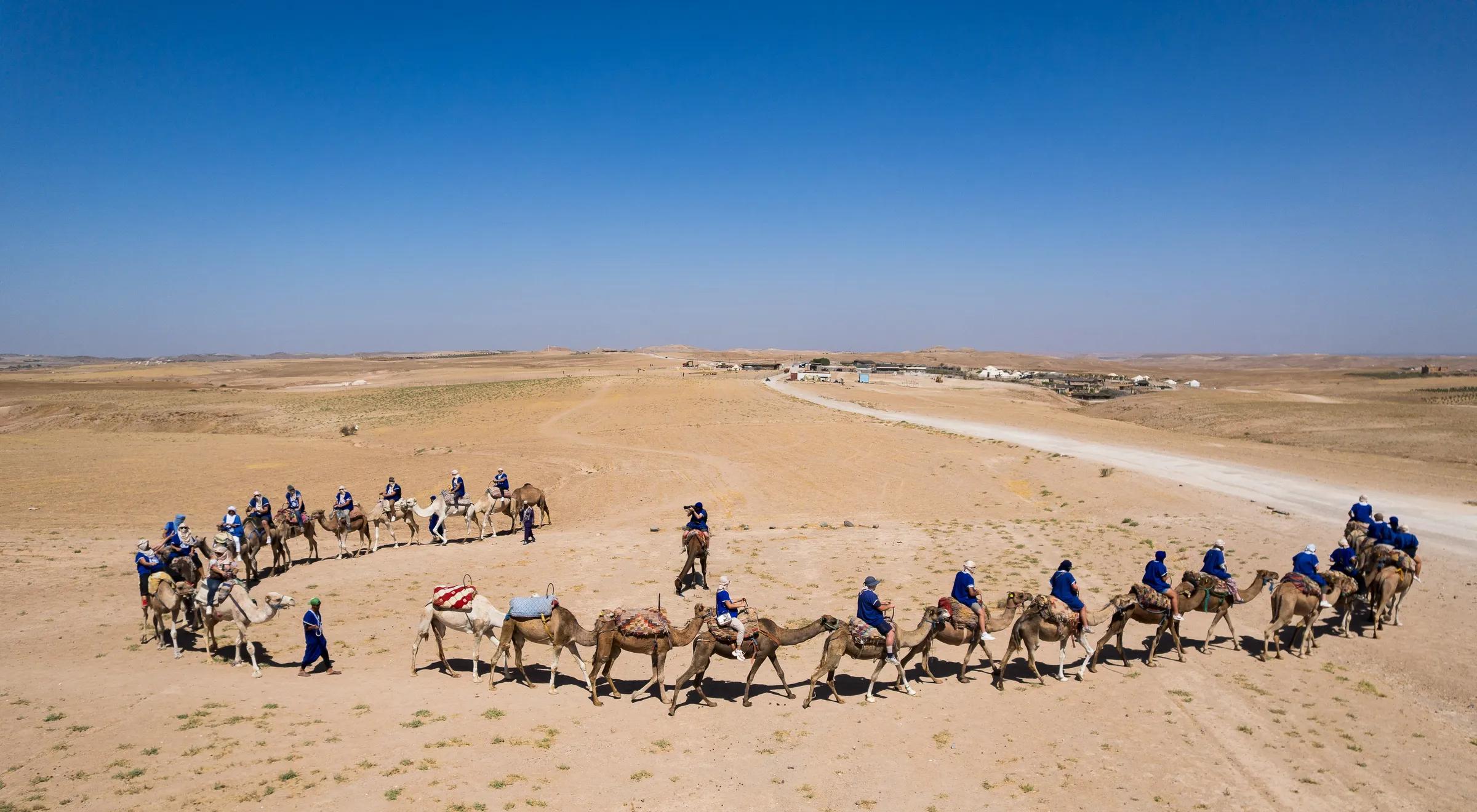 Vue du drone dans le désert d'Agafay : une caravane de dromadaires montés par les participants en gandoura bleue forme une boucle. Au centre, un photographe sur un dromadaire les photographie.